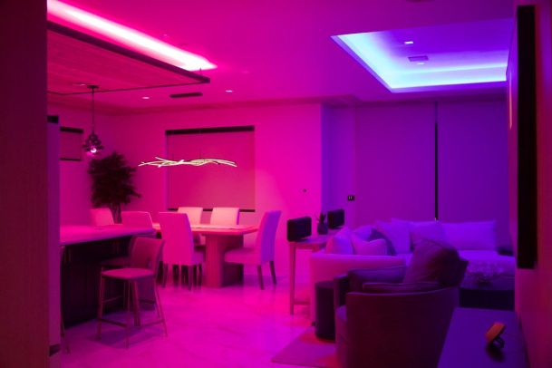 Living and dining area with pink and blue mood lighting, modern furniture, and a contemporary chandelier.