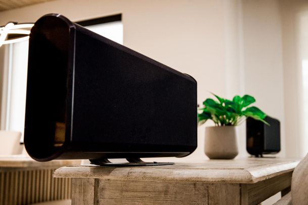 Side view of a sleek, modern speaker on a wooden table. The speaker is part of a sophisticated home audio setup.