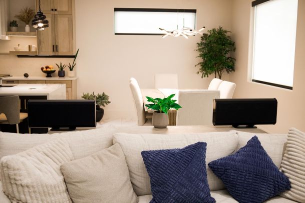 Living room with a light beige sofa, blue throw pillows, and a dining area in the background with modern pendant lights.