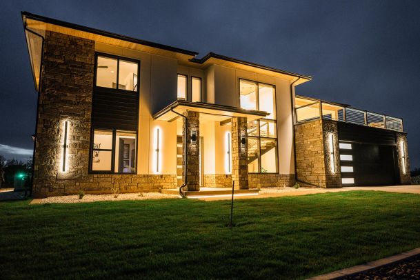 A contemporary house exterior at night, with illuminated stone pillars and modern vertical light fixtures highlighting the entrance and the side of the house.