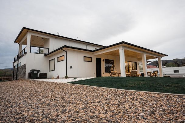 Modern two-story house with a spacious patio, gravel driveway, and a lush green lawn.