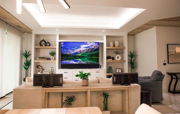 Living room with a large flat-screen TV mounted in a built-in wall unit. The room features a beige sofa, decorative shelves, and modern recessed lighting.