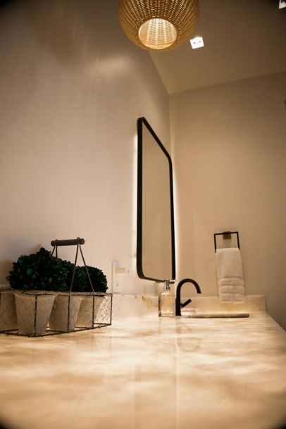 A bathroom with a modern vanity setup. It includes a backlit mirror, a wicker pendant light, a sleek black faucet, and a decorative plant in a wire basket on the countertop.