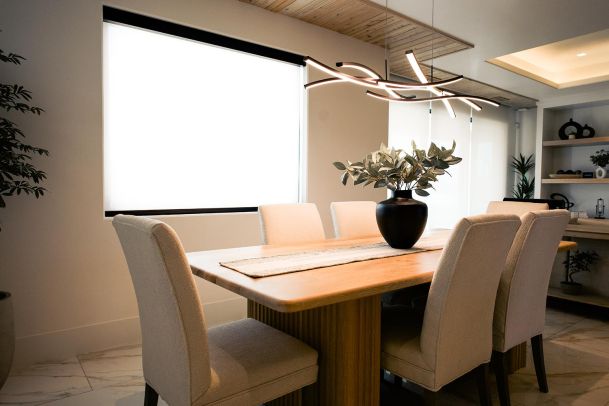 A dining area with a wooden table and six upholstered chairs. Above the table hangs a modern, sculptural light fixture. The room has large windows with white blinds and a plant in the corner.