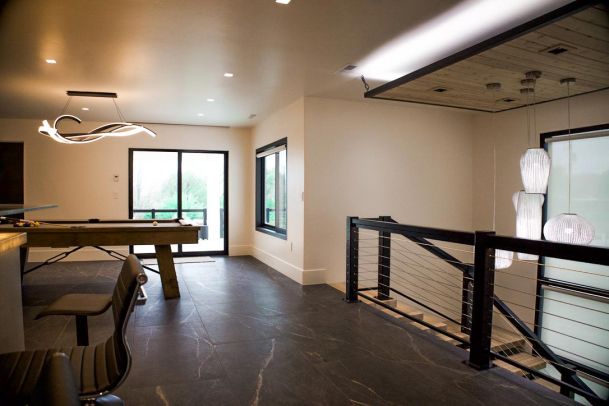 A modern game room with a pool table under a unique, sculptural light fixture. The room has large windows and a railing with cable wires leading to a staircase with hanging light fixtures.