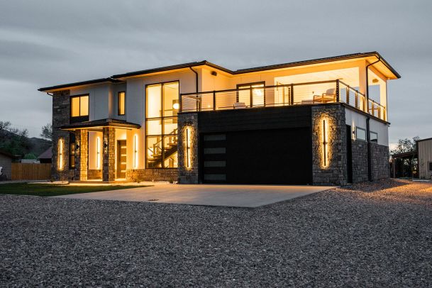 The exterior of a modern two-story house at dusk, featuring large windows and prominent vertical light fixtures on the stone facade. The house has a spacious driveway and landscaped front yard.