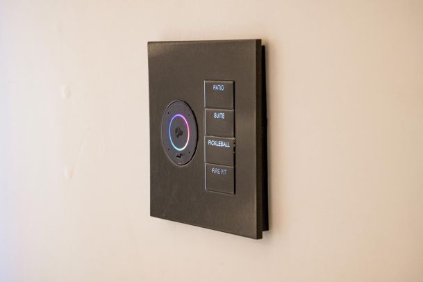 A black smart home control panel with buttons labeled for areas including Patio, Suite, Pickleball, and Fire Pit, featuring a Josh AI Nano control with a colored ring.