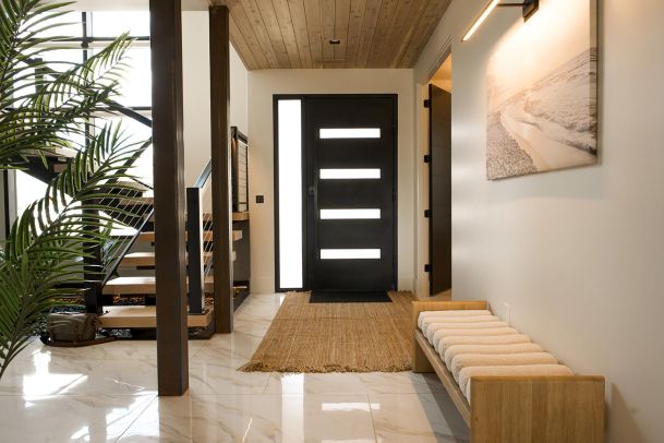 A modern entryway with a bench, plant, and staircase, featuring a wooden ceiling and a black front door with horizontal windows.