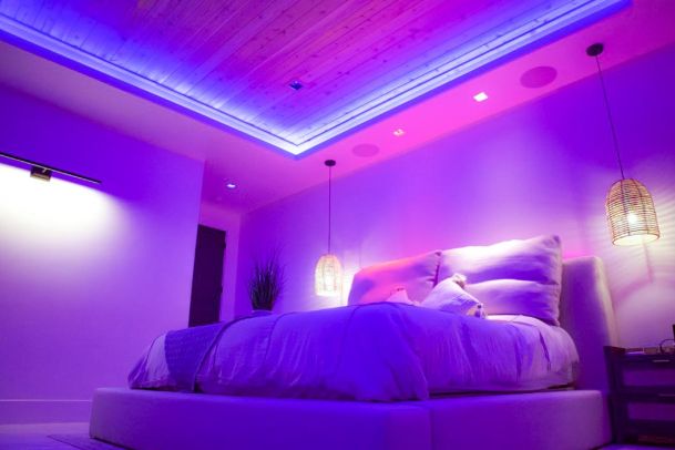 A bedroom illuminated with vibrant blue and pink lighting. The room features a large bed with a padded headboard, wooden ceiling with recessed lighting, and hanging woven pendant lights.