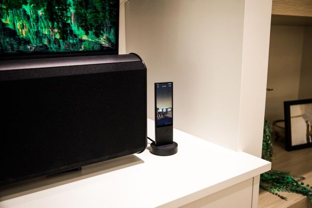 A remote control docked on a charging station beside a large speaker. The setup is part of a modern home entertainment system.