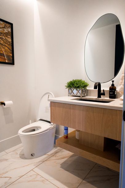 A clean, modern bathroom with a floating vanity, a uniquely shaped mirror, and a smart toilet. The vanity is decorated with a small potted plant and essential toiletries.